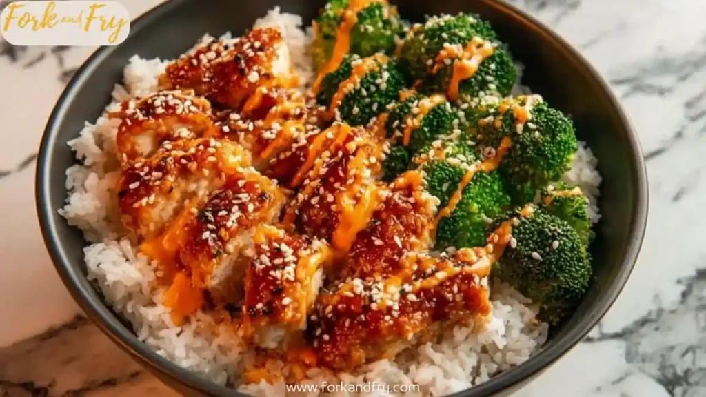 Bowl of crispy chicken over steamed rice and broccoli, topped with sesame seeds and spicy mayo drizzle