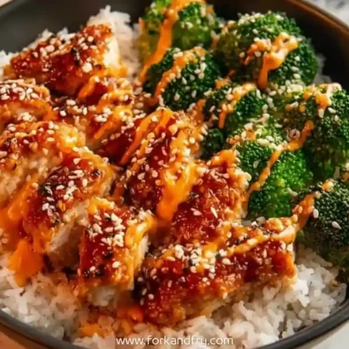 Bowl of crispy chicken over steamed rice and broccoli, topped with sesame seeds and spicy mayo drizzle