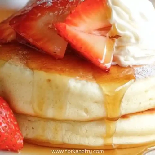 Stack of fluffy pancakes topped with sliced strawberries, whipped cream, and maple syrup drizzle