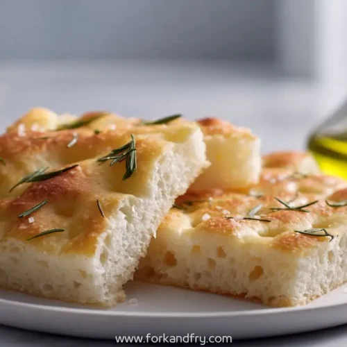 slices of golden focaccia bread with rosemary on a white plate next to olive oil