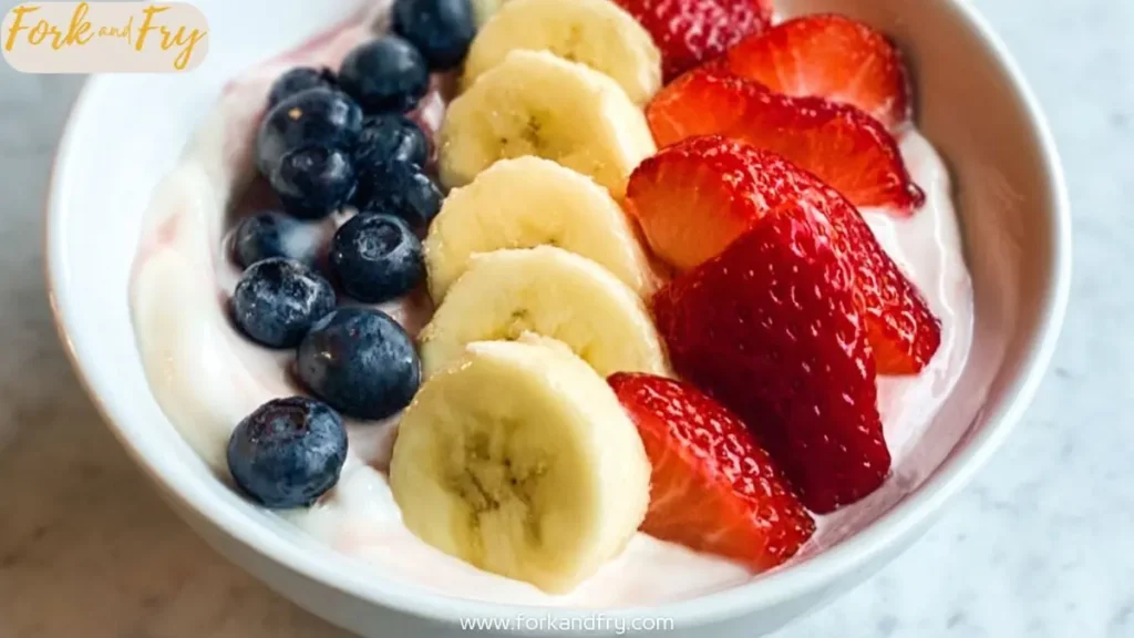Bowl of yogurt topped with fresh strawberries, banana slices, and blueberries in neat rows