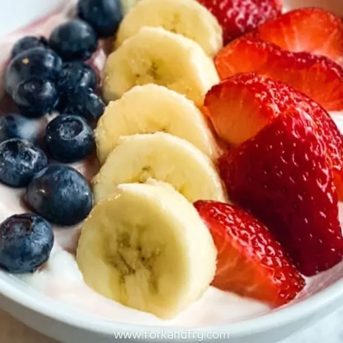 Bowl of yogurt topped with fresh strawberries, banana slices, and blueberries in neat rows