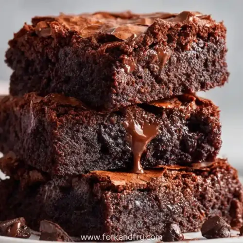 Stack of rich fudgy brownies with crackly tops on a white plate, melted chocolate drizzle, warm natural light.