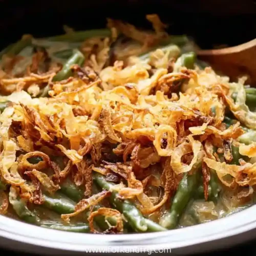 Green bean casserole topped with crispy fried onions in a slow cooker with a wooden spoon