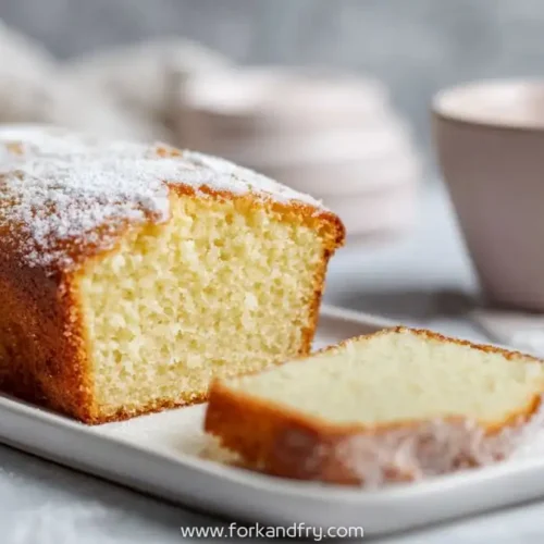 moist lemon loaf cake with powdered sugar on white plate next to pink coffee cup