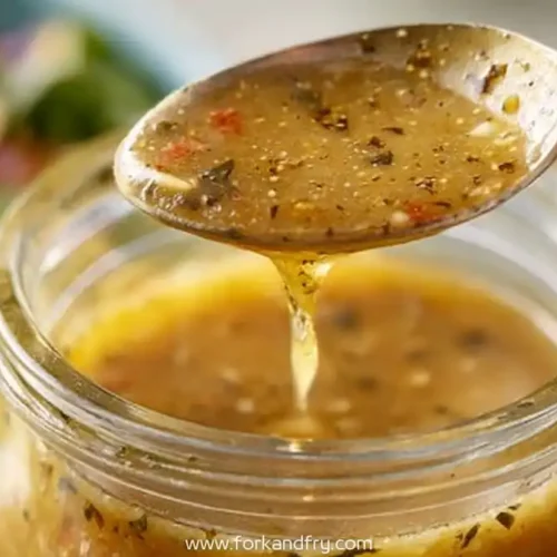 Spoon drizzling homemade healthy Italian dressing with herbs over a glass jar, salad in background
