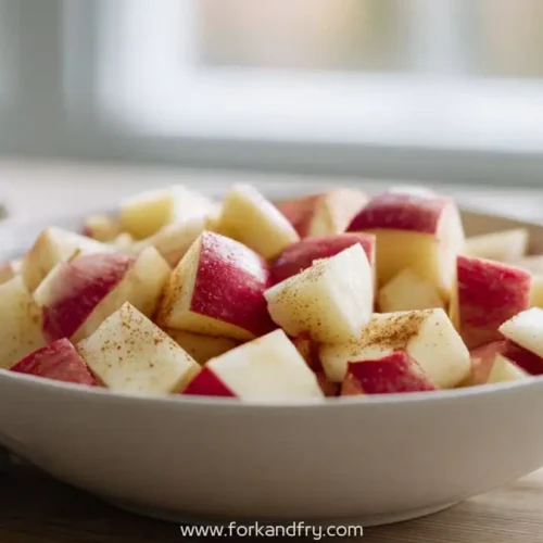 White bowl filled with red apple cubes dusted with cinnamon on wooden table in natural light