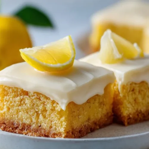 Moist lemon cake slice topped with creamy white frosting and fresh lemon wedge on plate